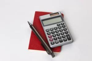 Calculatrice, carnet rouge et stylo noir posés sur une surface blanche.