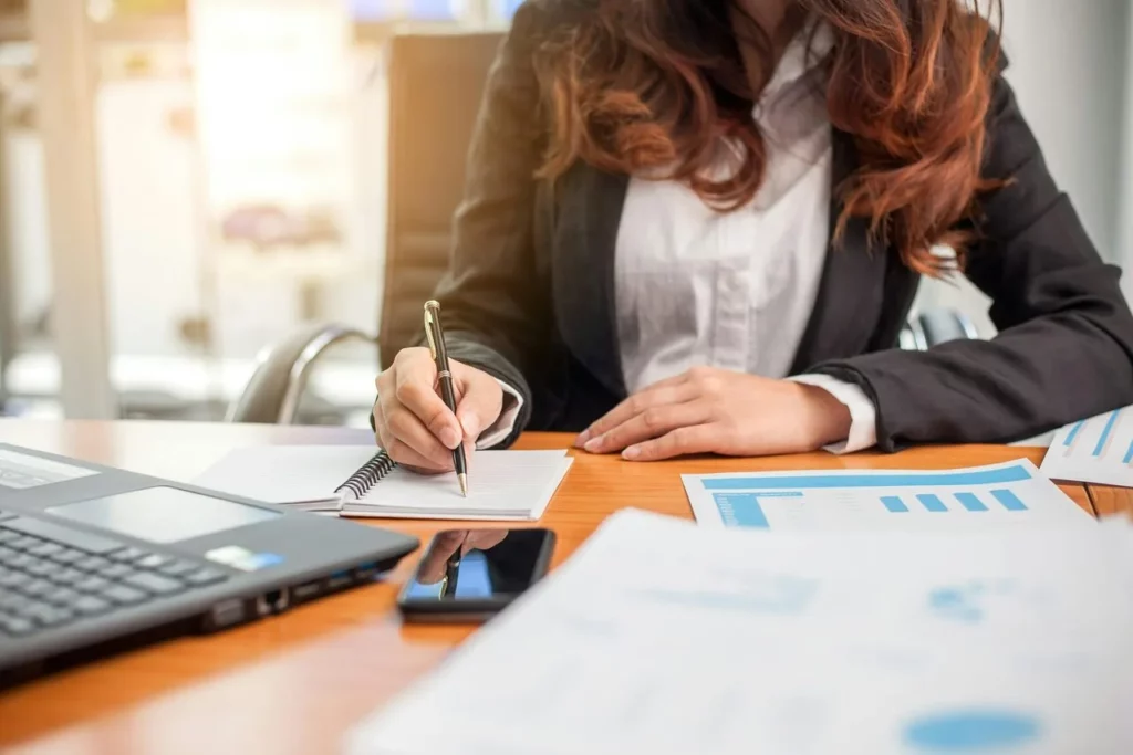 Une femme en costume écrivant dans un carnet à côté d'un ordinateur portable et de documents financiers, symbolisant l'assistance d'un expert-comptable pour les freelances.