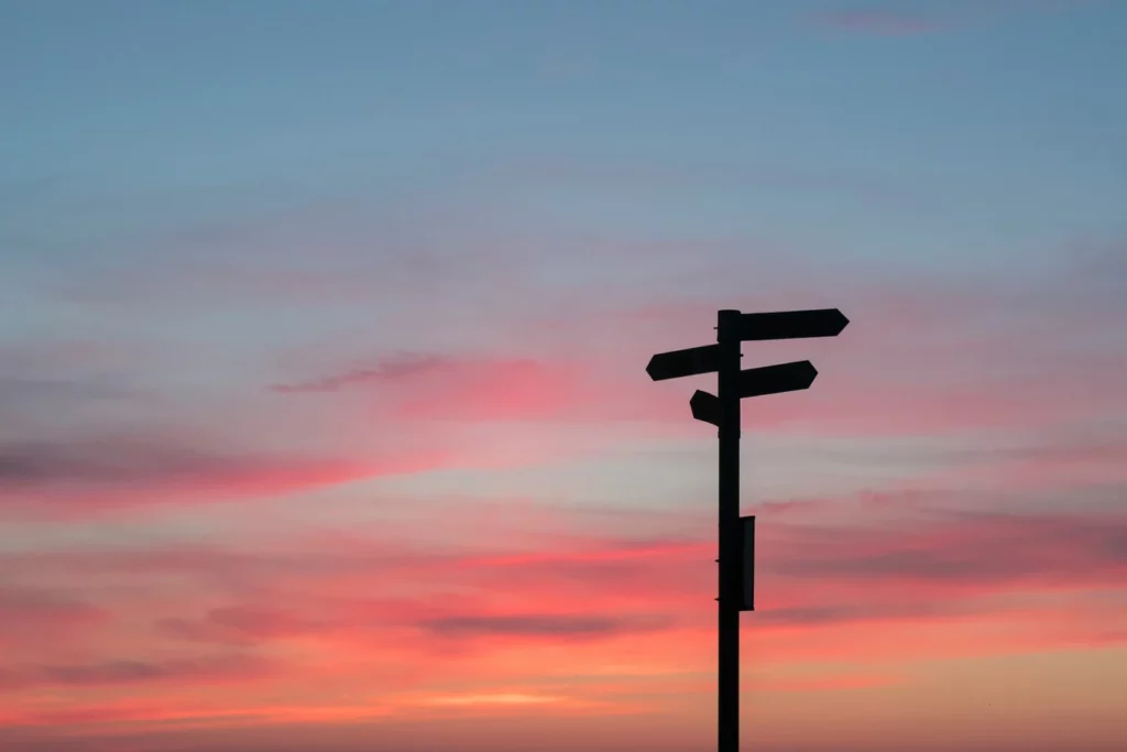 Panneau de direction silhouetté devant un ciel coloré au coucher du soleil, représentant les choix stratégiques d’un freelance pour créer une holding.